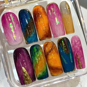 Puede incluir: Un juego de diez u&ntilde;as artificiales en una caja de pl&aacute;stico transparente. Cada u&ntilde;a presenta un dise&ntilde;o &uacute;nico con colores vibrantes como rosa, azul, naranja y verde, acentuado con ribetes dorados y l&aacute;mina iridiscente. Las u&ntilde;as tienen un acabado brillante.