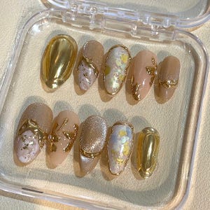 May include: A set of ten artificial nails in a clear plastic case. The nails feature a variety of designs, including solid gold, gold accents, and iridescent glitter. The base colors are neutral tones of beige and pink.