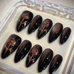 May include: A set of ten black, almond-shaped press-on nails. Each nail features a rose design in a metallic gold color. The nails are displayed in a clear plastic case.