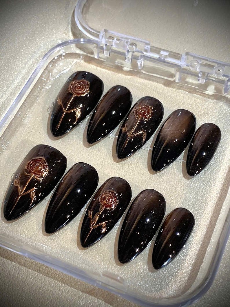 May include: A set of ten black, almond-shaped press-on nails. Each nail features a rose design in a metallic gold color. The nails are displayed in a clear plastic case.