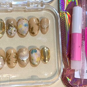 May include: A set of press-on nails in a clear plastic case. The nails are a light beige color with gold accents, glitter, and abstract designs. A pink nail file and nail glue are included.