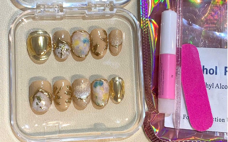 May include: A set of press-on nails in a clear plastic case. The nails are a light beige color with gold accents, glitter, and abstract designs. A pink nail file and nail glue are included.