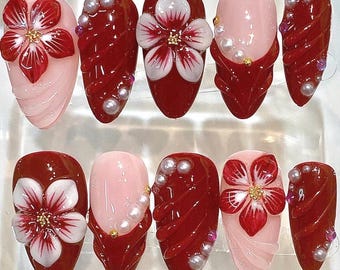 May include: A set of ten press-on nails featuring a glossy finish in deep red and soft pink. The designs include three-dimensional white and red floral accents, raised diagonal red ridges, and small white pearl embellishments with gold centers.