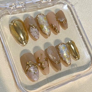 May include: A set of ten artificial nails in a clear plastic case. The nails feature a variety of designs, including gold metallic accents, glitter, and abstract patterns in neutral tones. The nails are oval-shaped.