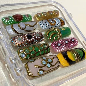 May include: A collection of ornate, decorative nail art designs in a clear plastic case. The nails feature various colors, including green, gold, pink, and white, with intricate patterns and embellishments. Some nails have a gold trim.
