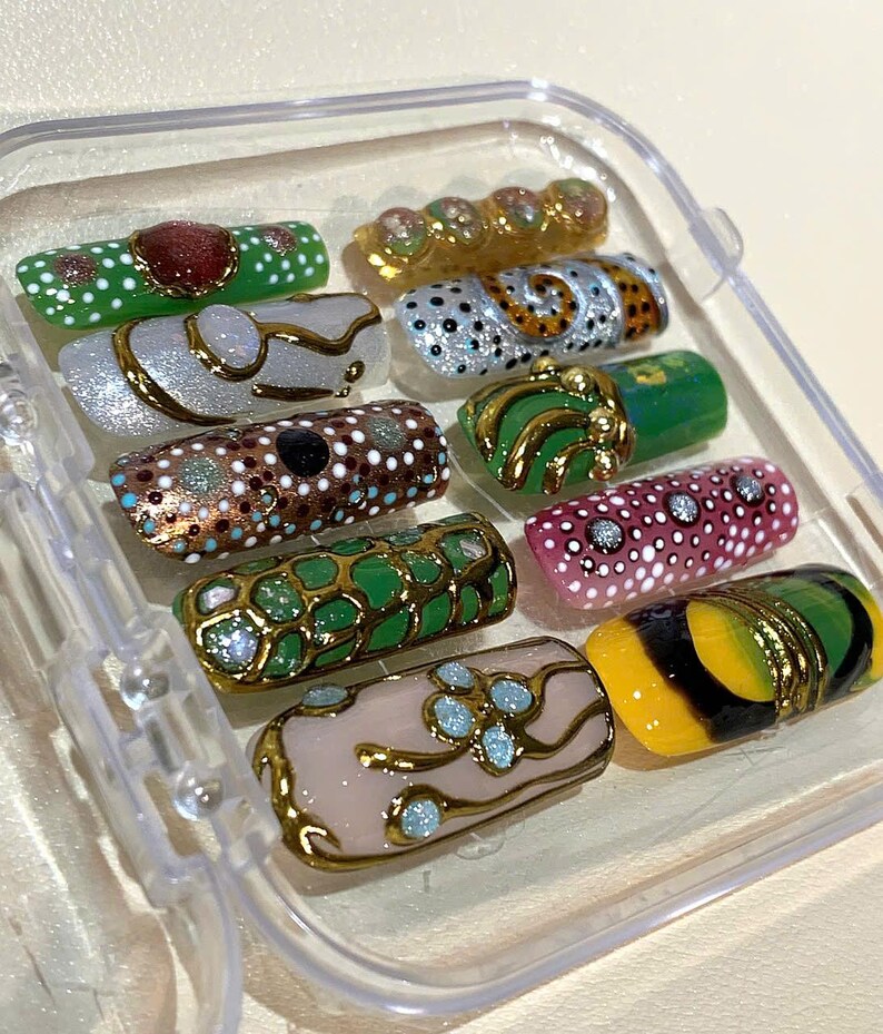 May include: A collection of ornate, decorative nail art designs in a clear plastic case. The nails feature various colors, including green, gold, pink, and white, with intricate patterns and embellishments. Some nails have a gold trim.