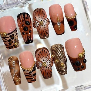 May include: A set of ten artificial nails with various designs. The nails feature a combination of pink, brown, and gold colors. Some nails have floral patterns, animal prints, and decorative embellishments. The nails are arranged in a clear plastic container.
