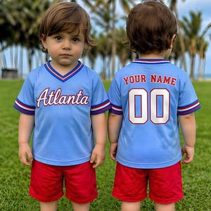 May include: Two young children wearing matching light blue baseball jerseys with "Atlanta" printed on the front and "YOUR NAME" and "00" on the back. They are also wearing red shorts and white slip-on shoes. The background features palm trees and a grassy field.