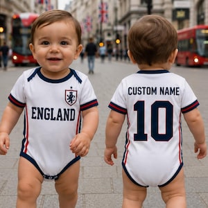 Könnte beinhalten: Zwei lächelnde Babys in weißen Bodysuits im England-Fußball-Stil. Auf der Vorderseite steht "ENGLAND" mit einem Wappen. Auf der Rückseite steht "CUSTOM NAME" und die Nummer "10". Beide tragen weiße Turnschuhe.