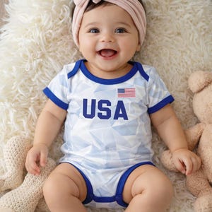 Puede incluir: Un beb&eacute; sonriente con un body blanco y azul con el tema de EE. UU. y una diadema rosa con lazo a juego. El body tiene ribetes azules y la bandera estadounidense. El beb&eacute; est&aacute; acostado sobre una alfombra blanca y esponjosa.