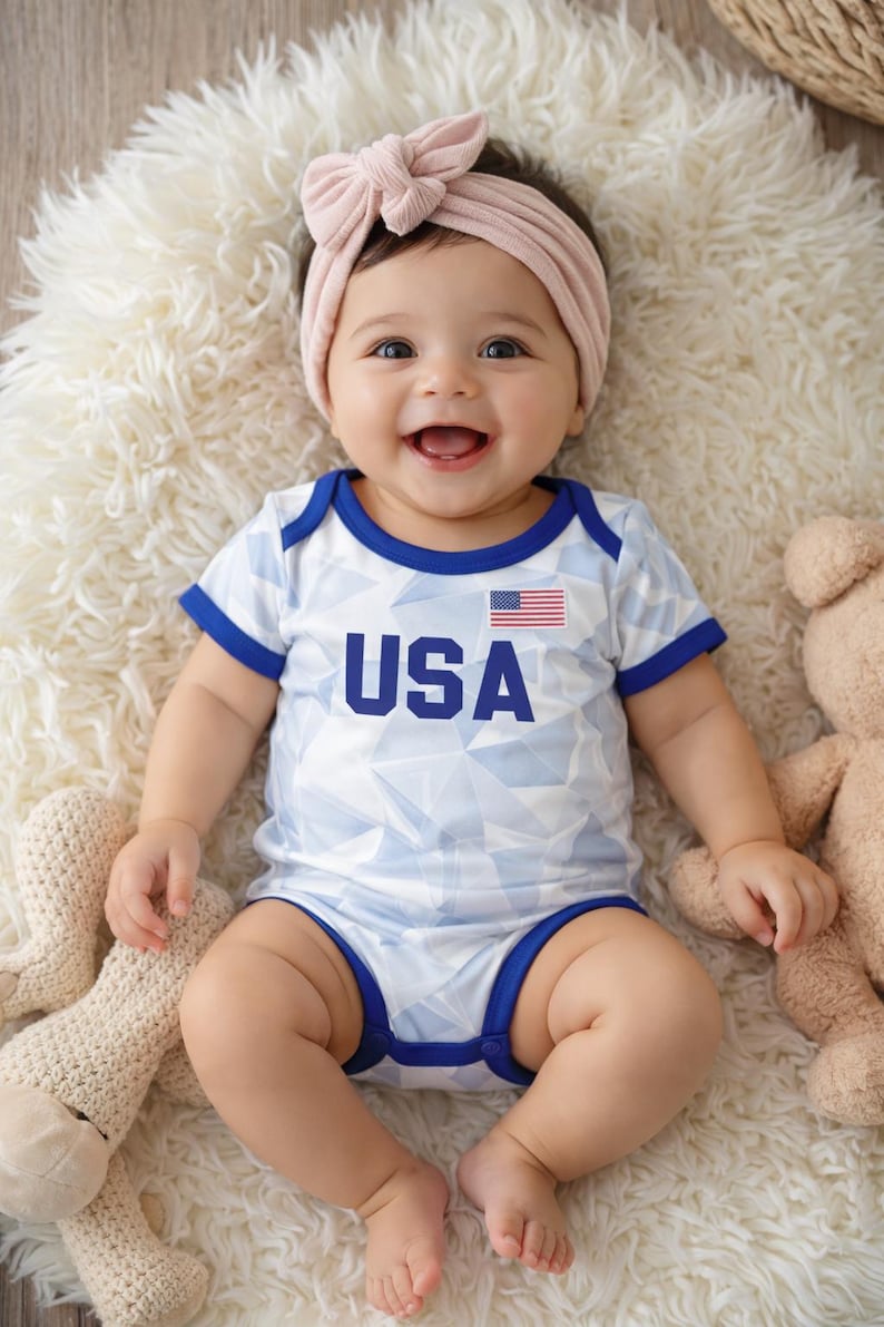 Puede incluir: Un beb&eacute; sonriente con un body blanco y azul con el tema de EE. UU. y una diadema rosa con lazo a juego. El body tiene ribetes azules y la bandera estadounidense. El beb&eacute; est&aacute; acostado sobre una alfombra blanca y esponjosa.