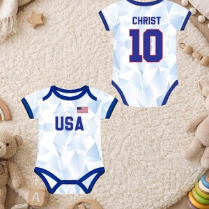 Puede incluir: Dos bodies blancos para beb&eacute; con ribetes azules y patrones geom&eacute;tricos. Uno tiene "USA" y una peque&ntilde;a bandera americana, el otro "CHRIST" y el n&uacute;mero "10" en azul. Los bodies se muestran sobre una alfombra cremosa y esponjosa.