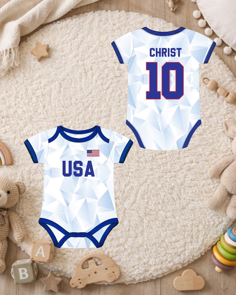 Puede incluir: Dos bodies blancos para beb&eacute; con ribetes azules y patrones geom&eacute;tricos. Uno tiene "USA" y una peque&ntilde;a bandera americana, el otro "CHRIST" y el n&uacute;mero "10" en azul. Los bodies se muestran sobre una alfombra cremosa y esponjosa.