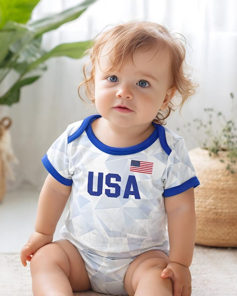 Puede incluir: Un beb&eacute; con un body blanco con un patr&oacute;n geom&eacute;trico, ribetes azules y la palabra "USA" en azul marino. La bandera estadounidense se muestra en la parte superior derecha. El beb&eacute; est&aacute; sentado frente a una planta.