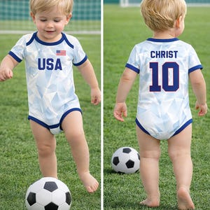 Puede incluir: Un beb&eacute; con un body de f&uacute;tbol azul claro y blanco con el texto "USA" y una bandera americana en la parte delantera. La parte trasera del body tiene el nombre "CHRIST" y el n&uacute;mero "10". El beb&eacute; est&aacute; de pie en un campo verde con un bal&oacute;n de f&uacute;tbol.