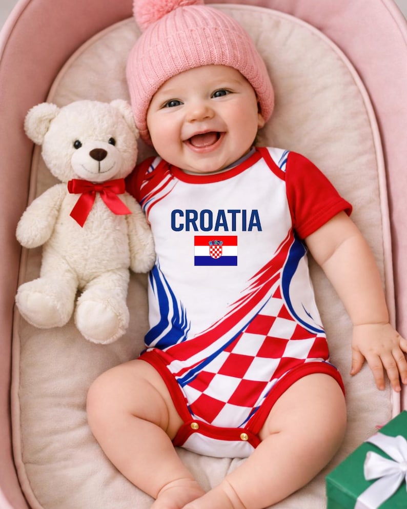 May include: A baby wearing a red, white, and blue bodysuit with the word "CROATIA" and the Croatian flag. The baby is also wearing a pink beanie and holding a white teddy bear with a red bow.