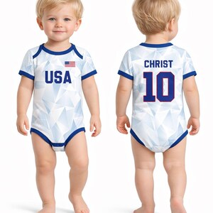 Puede incluir: Body de beb&eacute; blanco con un patr&oacute;n geom&eacute;trico y ribetes azules. La parte delantera presenta el texto "USA" y una peque&ntilde;a bandera estadounidense. La parte trasera muestra "CHRIST" y el n&uacute;mero "10" en azul.