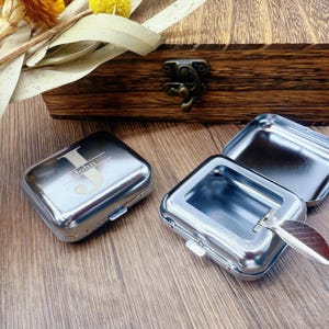 May include: Silver metal rectangular box with the name "John" engraved on the lid. The box is closed with a small clasp. Two other similar boxes are open, revealing their interior. A wooden box is in the background.
