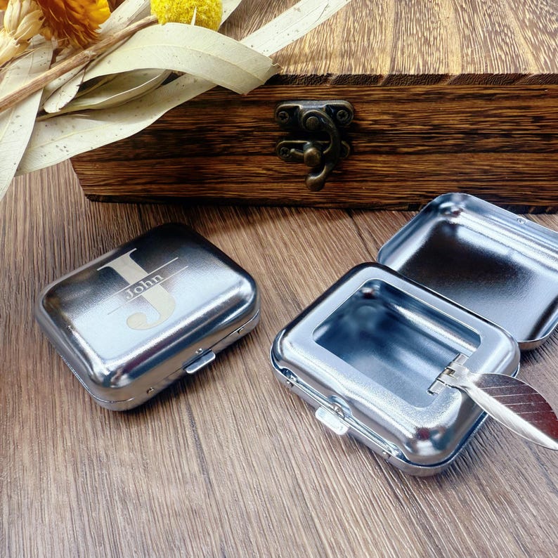May include: Silver metal rectangular box with the name "John" engraved on the lid. The box is closed with a small clasp. Two other similar boxes are open, revealing their interior. A wooden box is in the background.
