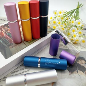 Puede incluir: Varios atomizadores de perfume de colores, rosa, dorado, rojo y negro, con detalles plateados. Un atomizador azul está grabado con el nombre "Daniela". También hay un atomizador de vidrio transparente y un ramo de flores blancas.