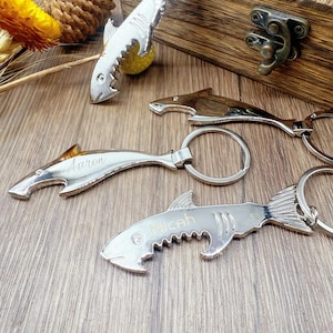 Könnte beinhalten: Haiförmige Flaschenöffner und Schlüsselanhänger in Silber- und Holztönen. Die silbernen Öffner sind mit den Namen "Aaron" und "Micah" graviert. Die Artikel sind auf einer Holzoberfläche angeordnet.