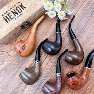 May include: An assortment of wooden tobacco pipes in various shades of brown, with black stems and silver accents. One pipe is inside a wooden box with the text "GROOMSMAN HENOK JULY 28, 2023". White flowers are in the background.