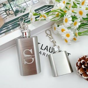 Op de afbeelding: Twee roestvrijstalen heupflacons met sleutelhangers. Op één flacon staat de letter "S" en de naam "Stefan" gegraveerd. De andere flacon is effen. De flacons zijn rechthoekig met een zilveren dop en sleutelhanger.