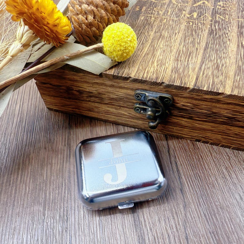 May include: A silver square metal box with the letter "J" and the name "John" engraved on the top. The box is next to a wooden box with a metal clasp and decorative dried flowers.