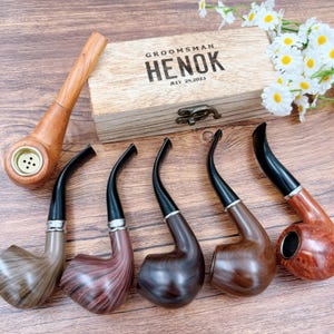 May include: An assortment of wooden tobacco pipes in various shades of brown, with black stems and silver accents. A wooden box with the text "GROOMSMAN HENOK JULY 28, 2023" is also present. The pipes are arranged on a wooden surface.