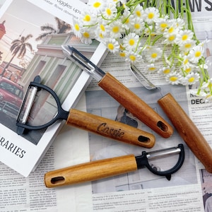 May include: A collection of kitchen peelers with wooden handles and stainless steel blades. One peeler has the name "Cassie" engraved on the handle. The peelers are arranged on a newspaper background with white flowers.