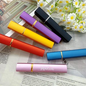 May include: A collection of colorful, refillable perfume atomizers. The atomizers are in various colors, including red, yellow, purple, black, and blue. One pink atomizer has the name "Malia" in cursive. Each atomizer has a gold-colored band.