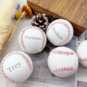May include: White baseballs with red stitching, each bearing a name: Paxton, Beckett, and Trey. The baseballs are displayed on a newspaper background, alongside a pinecone and dried flowers.