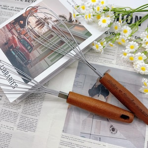 Puede incluir: Dos batidores de acero inoxidable con mangos de madera. Los mangos están grabados con los nombres "Kristen & Brandon" y la fecha "10.18.2023". Los batidores están sobre un periódico y cerca de flores blancas.