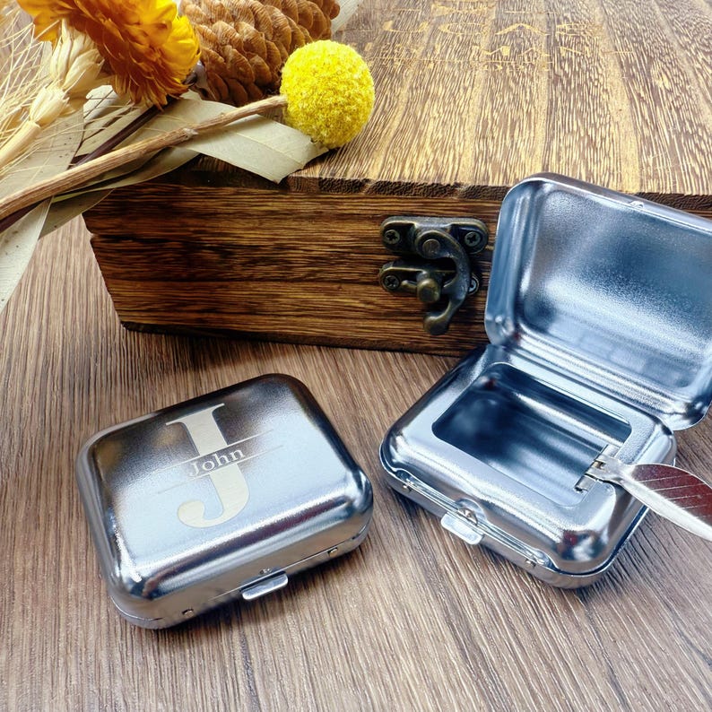 May include: Two small, shiny, silver metal boxes, one open and one closed, sit on a wooden surface. The closed box has the letter "J" and the name "John" engraved on it. A wooden box with a metal clasp is in the background.