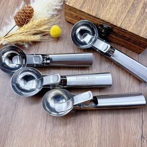 May include: Four stainless steel ice cream scoops with a trigger mechanism, arranged on a wooden surface. One scoop is engraved with the name "Ryan Kile" and the word "Groomsman". The scoops are a shiny silver colour.