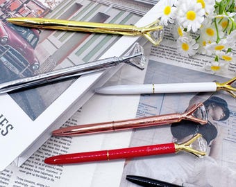 May include: An assortment of elegant pens with diamond-shaped tops. The pens come in gold, silver, rose gold, red, white, and black. The pens are arranged on a newspaper background with a bouquet of white flowers.