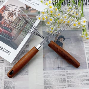 Puede incluir: Dos batidores de acero inoxidable con mangos de madera. Los mangos están grabados con los nombres "Kristen & Brandon" y la fecha "10.15.2023". Los batidores están sobre un periódico con flores blancas en el fondo.