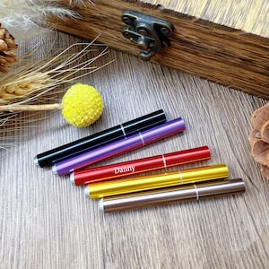 May include: A collection of six slim, metallic pen-style containers in various colors: black, purple, red, gold, and brown. The word "Danny" is printed on the red pen. The pens are arranged on a wooden surface.