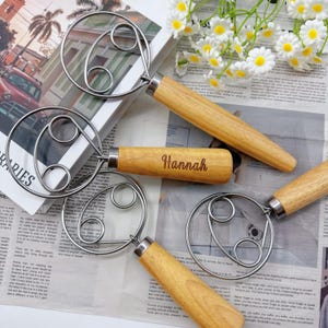 Puede incluir: Varios batidores de acero inoxidable con mangos de madera se exhiben. Un mango está grabado con el nombre "Hannah". Los batidores tienen un diseño circular único. El fondo incluye un libro, un periódico y flores blancas.