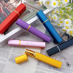 Op de afbeelding: Een verzameling kleurrijke, rechthoekige parfumverstuivers in rood, paars, blauw, roze, geel en marineblauw. Elke verstuiver heeft een goudkleurige band en dop. De roze verstuiver heeft de naam "Malia".
