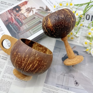 Puede incluir: Dos tazas artesanales de cáscara de coco. Una taza tiene un asa y una base de madera. La otra taza está sobre un tallo de madera. La palabra "Myles" está grabada en el lateral. Sobre un fondo de periódico.