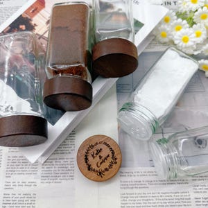 Op de afbeelding: Transparante glazen kruidenpotten met donkere houten deksels. Eén pot bevat een bruin poeder, een andere is gevuld met wit zout. Een houten deksel met de tekst "Katie & Connor 2023" is ook zichtbaar. De potten staan op een bedrukt oppervlak.