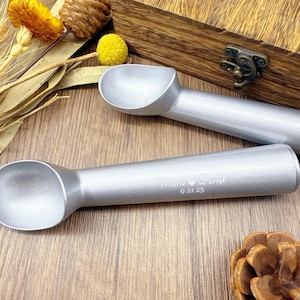 May include: Two silver ice cream scoops with engraved text reading "Marie Darryl" and the date "9 21 25". The scoops are on a wooden surface, with decorative dried flowers and a wooden box in the background. The scoops are made of metal.