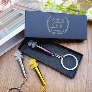 以下が含まれることがあります： 銀、金、虹色のミニチュアマイクロフォンキーホルダーのセット。1つのキーホルダーは、「Chris」という名前と「Groomsman 9.13.2026」というテキストが入ったギフトボックスに入っています。キーホルダーは木製の表面にあります。