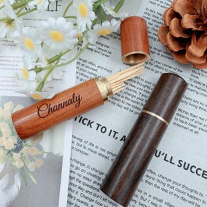 May include: Two wooden toothpick holders. One is open, revealing toothpicks, and engraved with "Channaly". The other is closed. Both have brass accents. The holders are on a background of white flowers and printed paper.