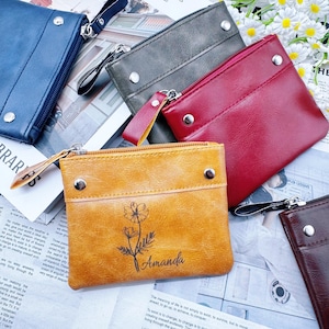 Op de afbeelding: Een verzameling kleine, met rits afsluitbare portemonnees in verschillende kleuren: blauw, grijs, rood en bruin. De voorste portemonnee is bruin met een bloemenontwerp en de naam "Amanda". Elke portemonnee heeft een zilveren rits.