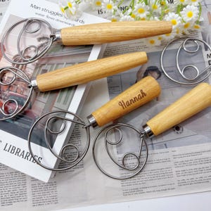 Puede incluir: Varios batidores de masa de acero inoxidable con mangos de madera están colocados sobre un periódico. Un mango está grabado con el nombre "Hannah". Los batidores tienen un diseño circular y están hechos de metal y madera.