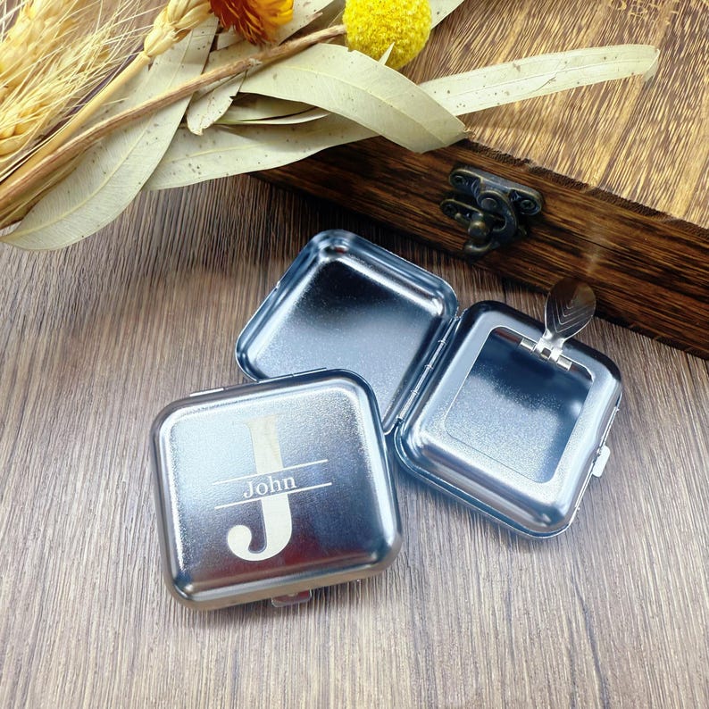 May include: Three open, square, silver-colored metal boxes with hinged lids. One box has the name "John" and a stylized "J" engraved on the lid. The boxes are on a wooden surface, with a wooden box and dried flowers in the background.