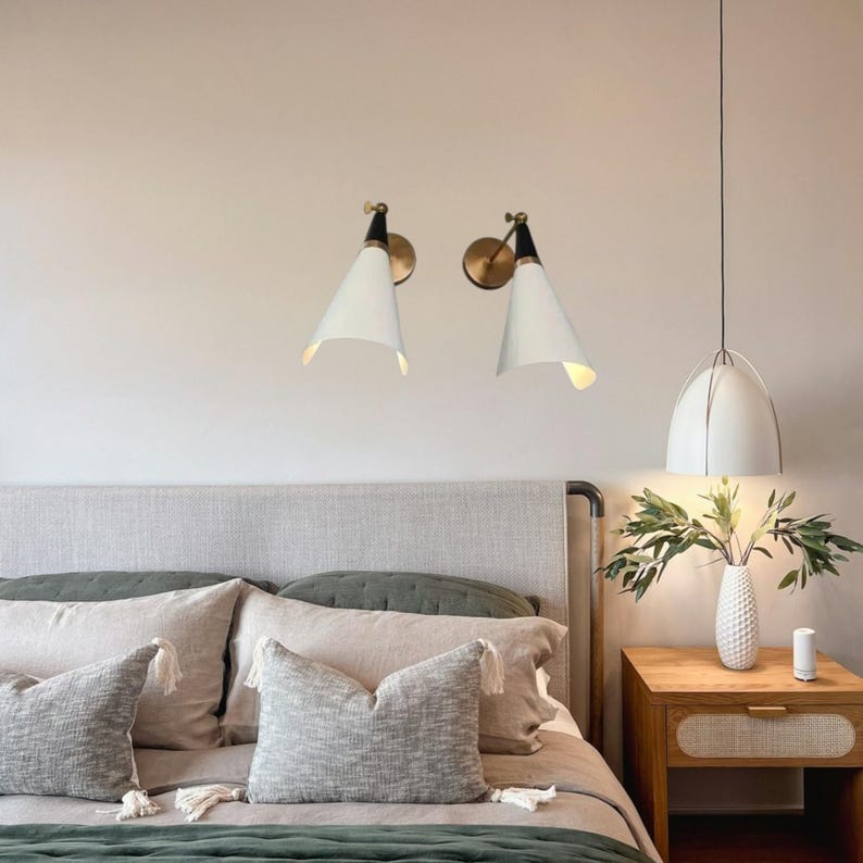 May include: Two white cone-shaped wall sconces with brass accents and a black top are mounted on a light-colored wall. A white pendant light hangs from the ceiling. The scene includes a bed with pillows and a wooden nightstand.