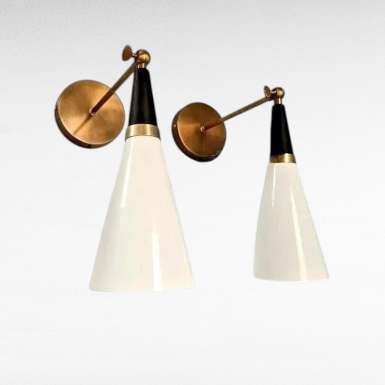 May include: Two mid-century modern wall sconces. Each sconce features a white, cone-shaped shade, a black top, and a brass-colored mounting plate and arm. The sconces are designed for wall mounting and provide a stylish lighting solution.